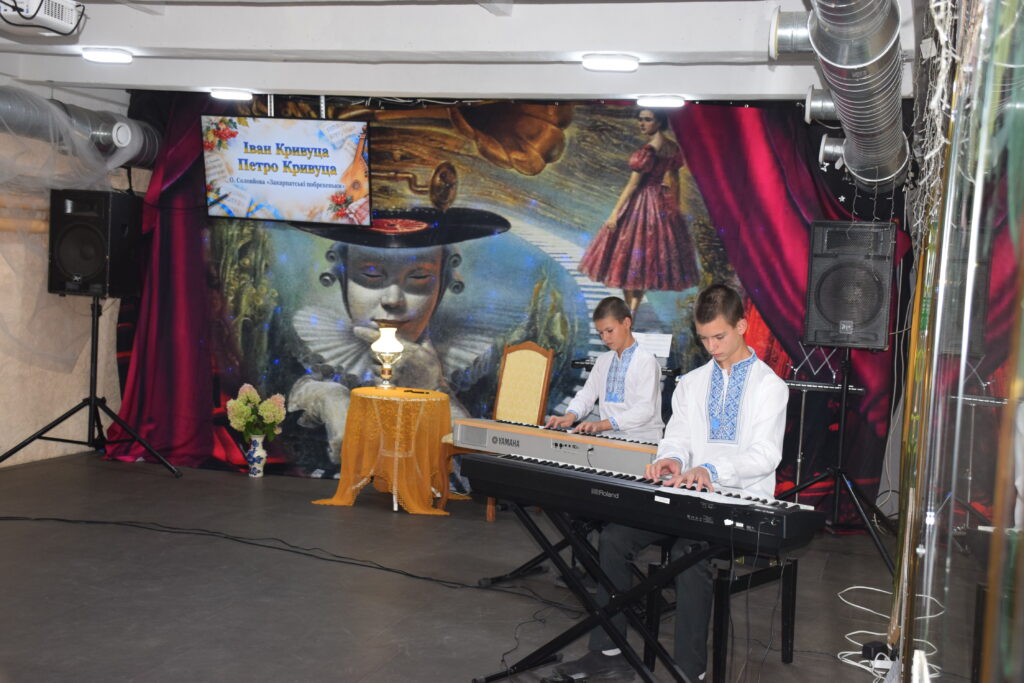 Двоє хлопців у вишиванках грають на електронних піаніно Yamaha та Roland на сцені, де на банері вказано імена Іван Кривуца та Петро Кривуца.