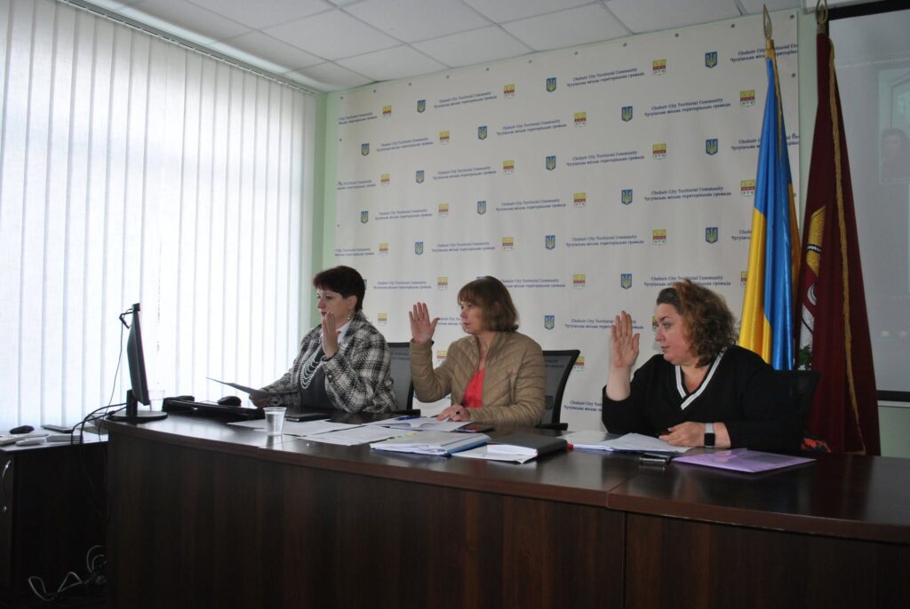Три жінки сидять за столом під час офіційної зустрічі, піднімаючи руки, на тлі банера з написом 