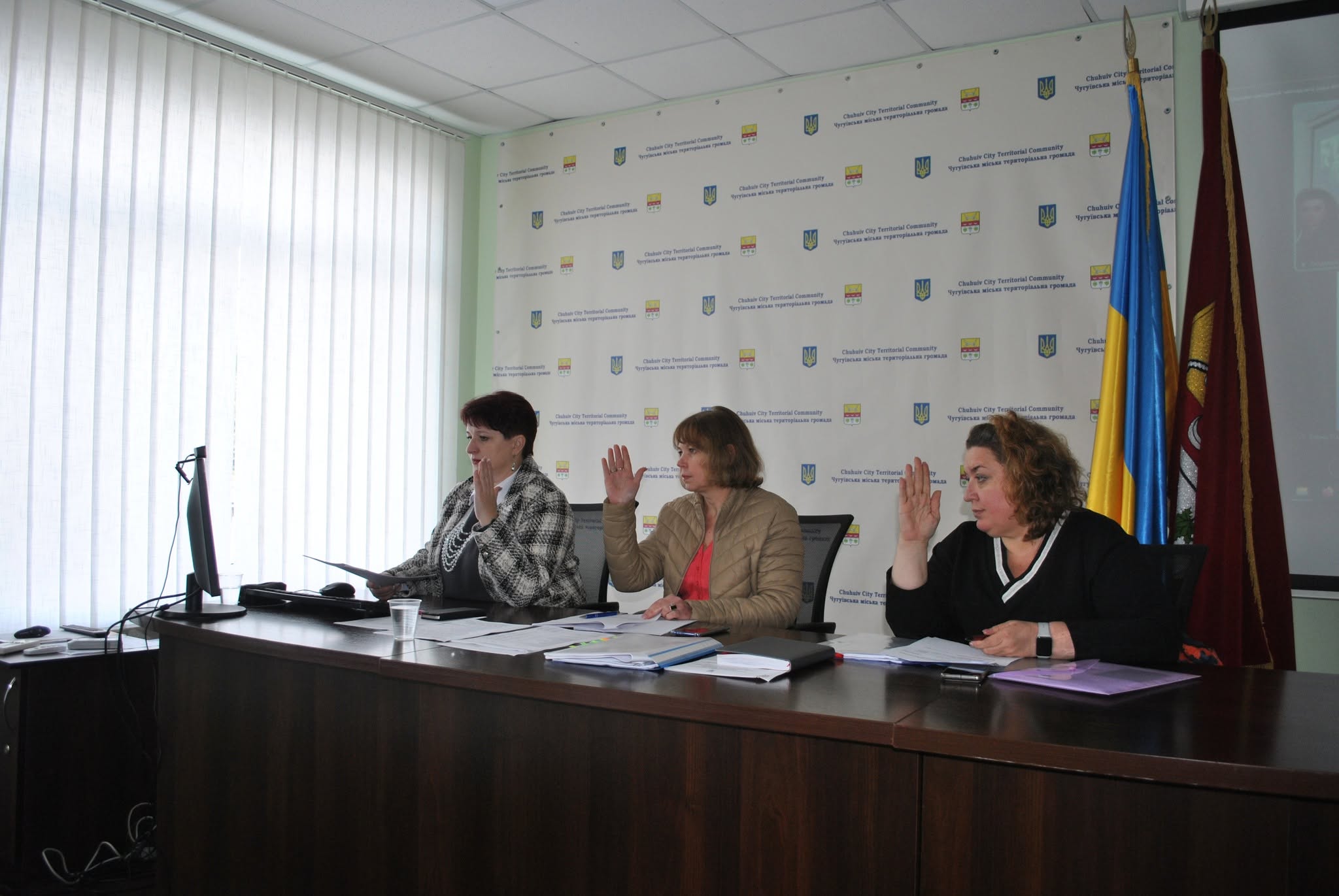 Три жінки сидять за столом під час офіційної зустрічі, піднімаючи руки, на тлі банера з написом 