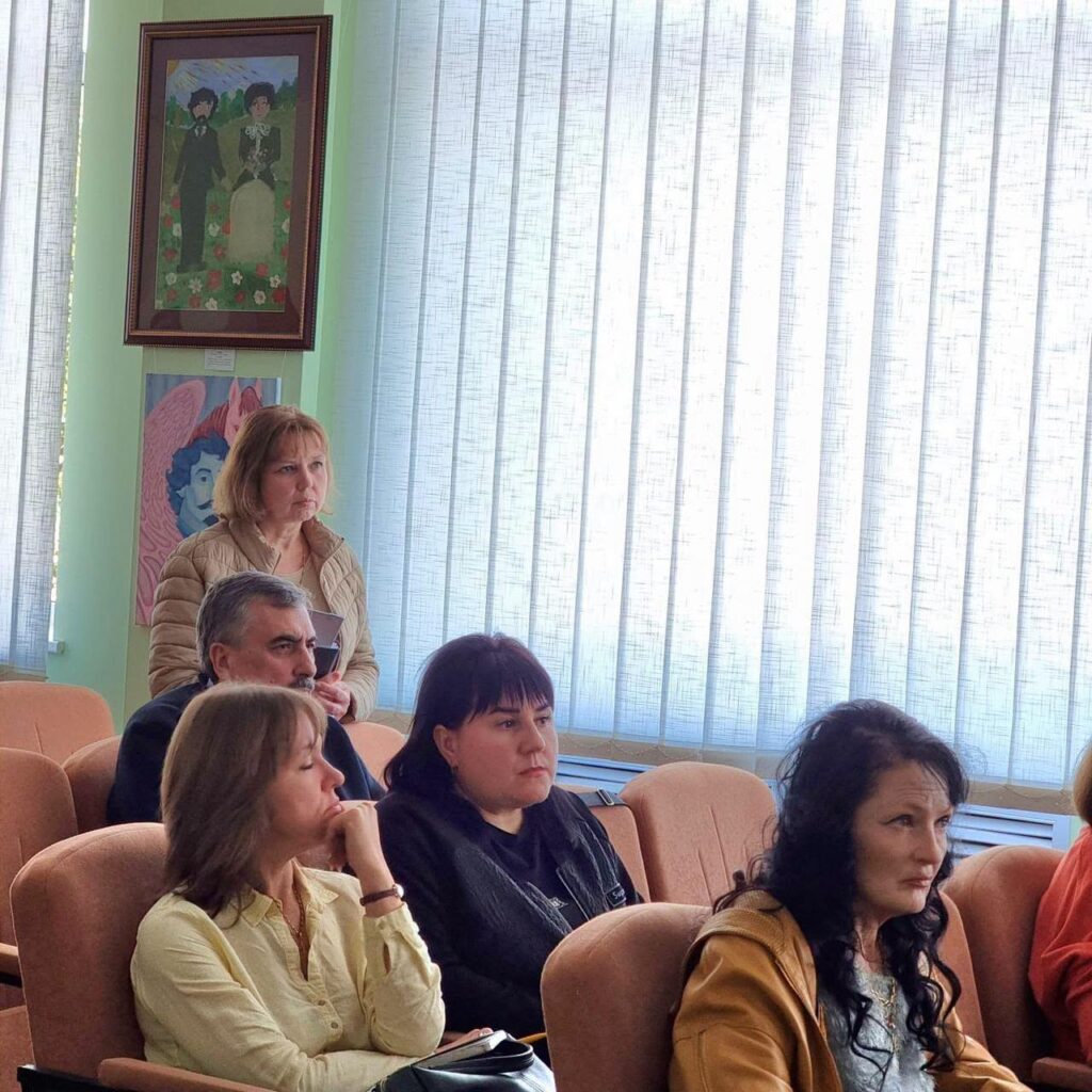 Група людей сидить у залі з вікнами, вкритими вертикальними жалюзі, та стінами, прикрашеними художніми полотнами.