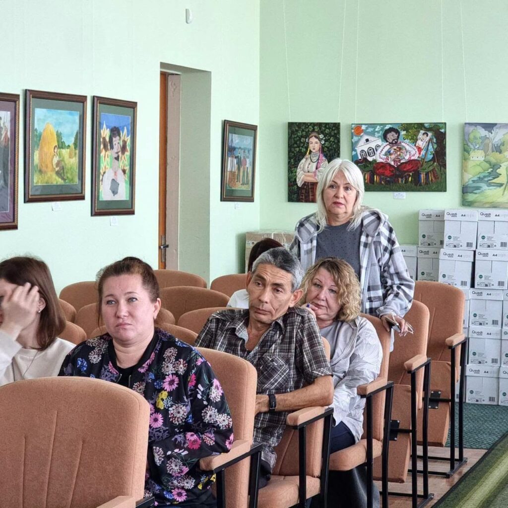 Група людей сидить у залі з рядами крісел, на стінах за ними висять художні полотна в рамах.