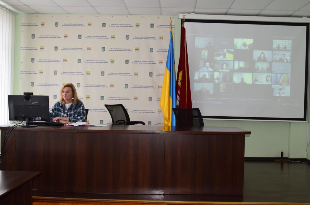 Жінка сидить за столом під час відеоконференції на тлі банера з написом