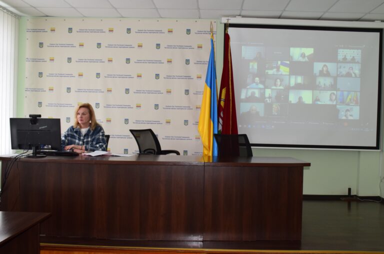Жінка сидить за столом під час відеоконференції на тлі банера з написом 