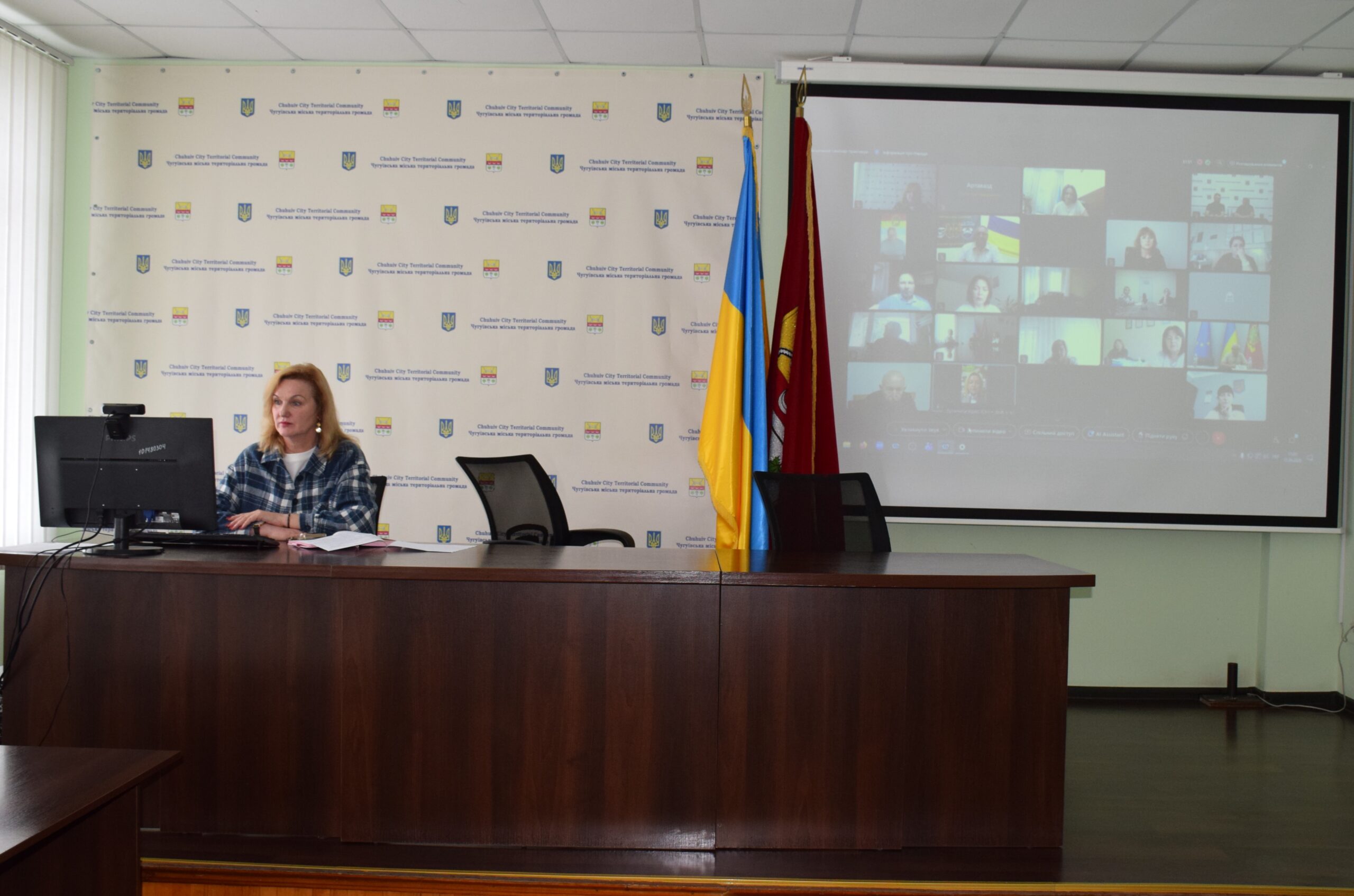 Жінка сидить за столом під час відеоконференції на тлі банера з написом 