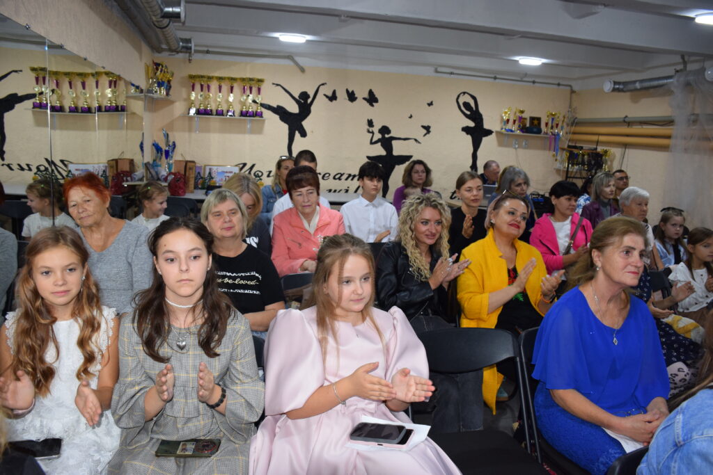 Група людей, переважно дітей та дорослих, сидить у залі та плескає в долоні, а на стіні позаду розміщені трофеї та силуети балерин.