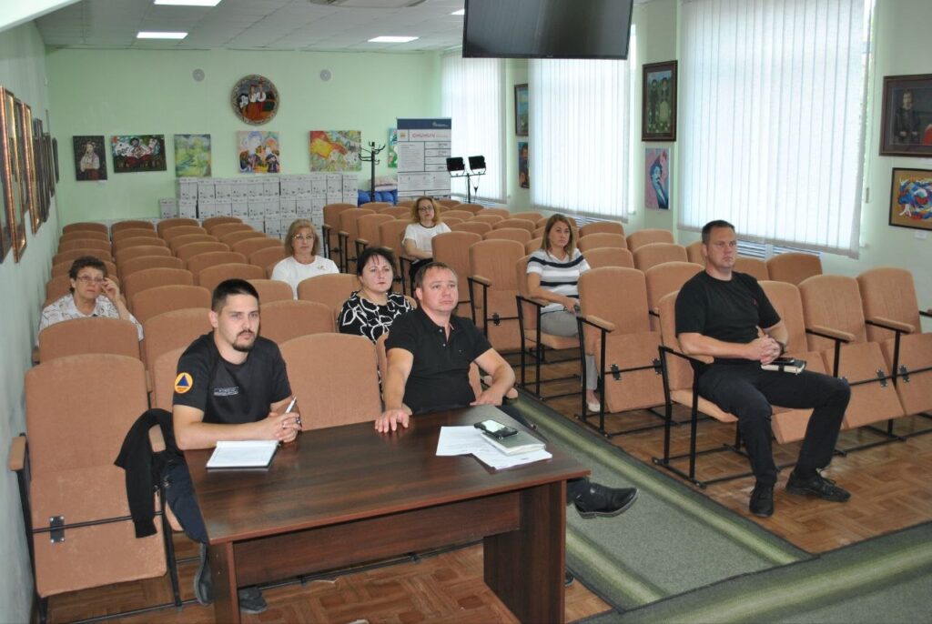 Учасники засідання сидять у залі з рядами крісел, а на передньому плані чоловіки в чорному одязі сидять за столом з документами.