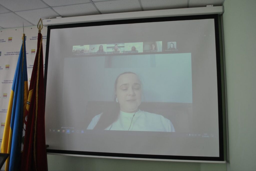Проекційний екран відображає відеоконференцію з учасниками, поруч стоять прапори.