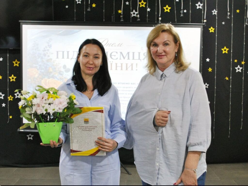 Жінка тримає букет квітів та грамоту від Національної місії територіальної громади на тлі банера з написом «Підсумки переможці».