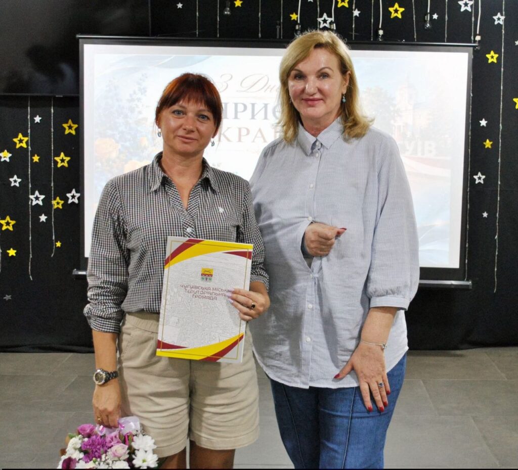Жінка тримає грамоту від Чугуївської міської територіальної громади та букет квітів, стоячи поруч з іншою жінкою на тлі банера зі словами «Україна».
