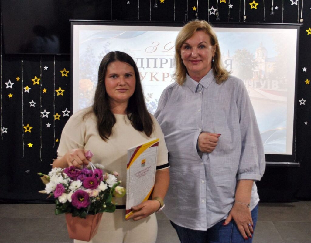 Дві жінки тримають квіти та грамоту на тлі банера з написом «З Днем вчителя в Україні».