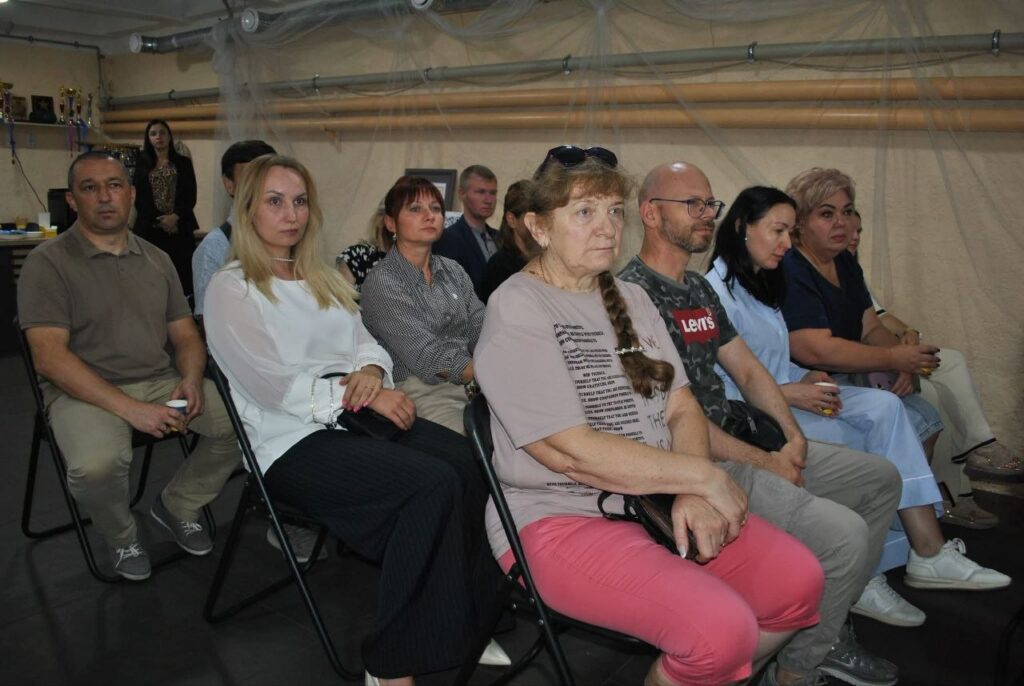 Група людей сидить на складних стільцях у залі, уважно слухаючи виступ.