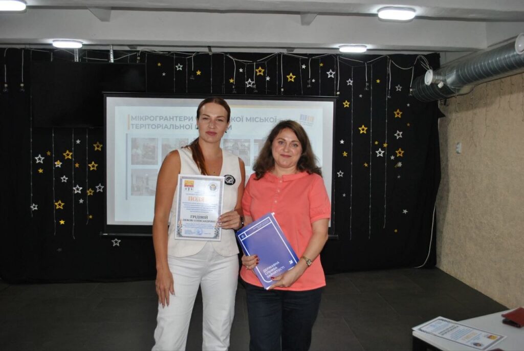 Дві жінки тримають сертифікат та синю папку з написом «Програма служби спільноти» перед екраном із текстом «Мікрогранти територіальної громади».