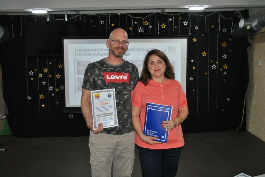 Чоловік і жінка тримають нагородні документи, зокрема подяку та синю книжку Державної служби зайнятості, на тлі банера з написом «Мікрогранти».