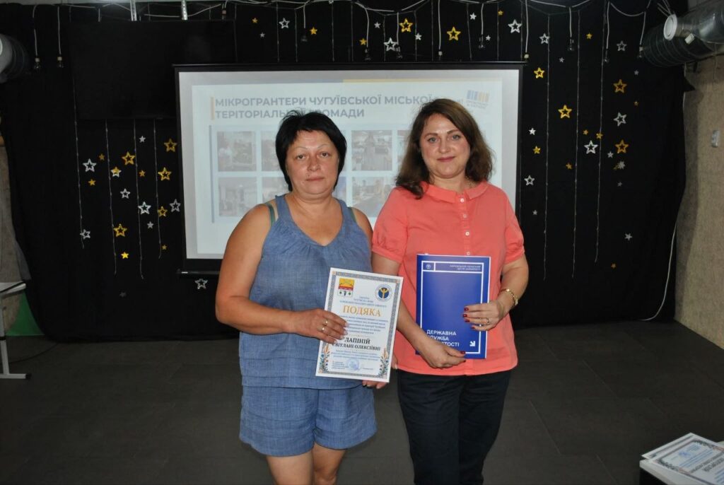 Дві жінки тримають документи «Подяка» та «Державна служба якості» перед банером з написом «Мікрогрантери Чугуївської міської територіальної громади».