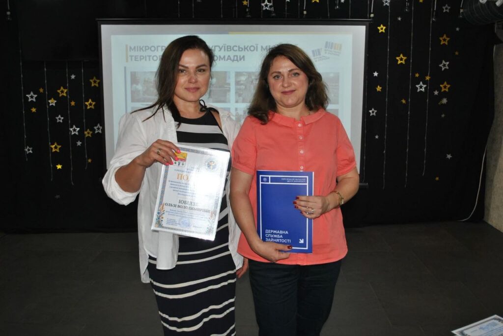 Дві жінки тримають у руках документи, зокрема державну службу зайнятості, на тлі банера з написом «Мікрогрупової територіальної громади».