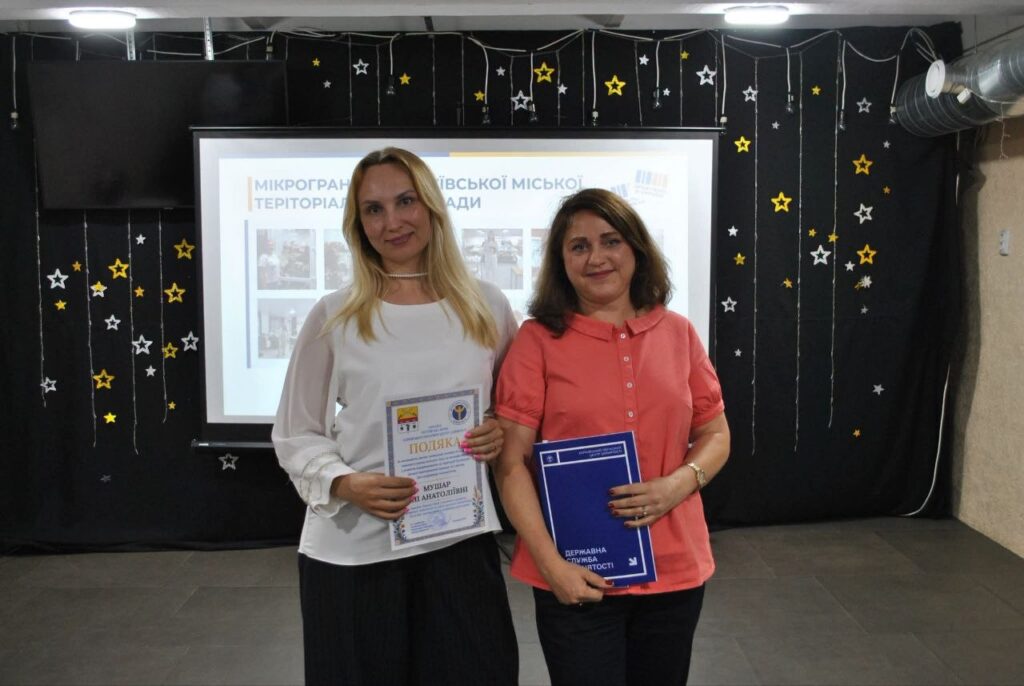 Дві жінки тримають нагороду «Подяка» та синю книжку «Державна служба зайнятості» перед екраном із написом «Мікрографія Територіальної громади».