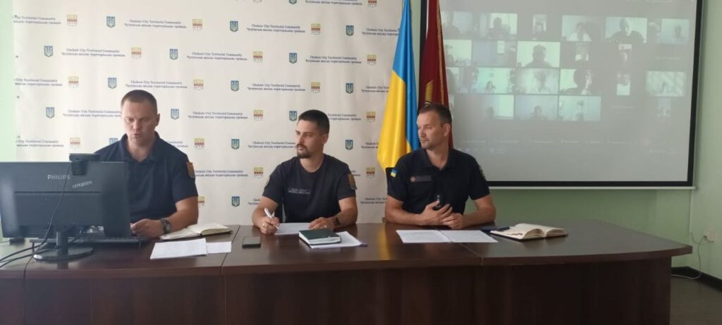 Три чоловіки в формі сидять за столом під час відеоконференції з проєкцією учасників на екрані та прапорами на тлі.
