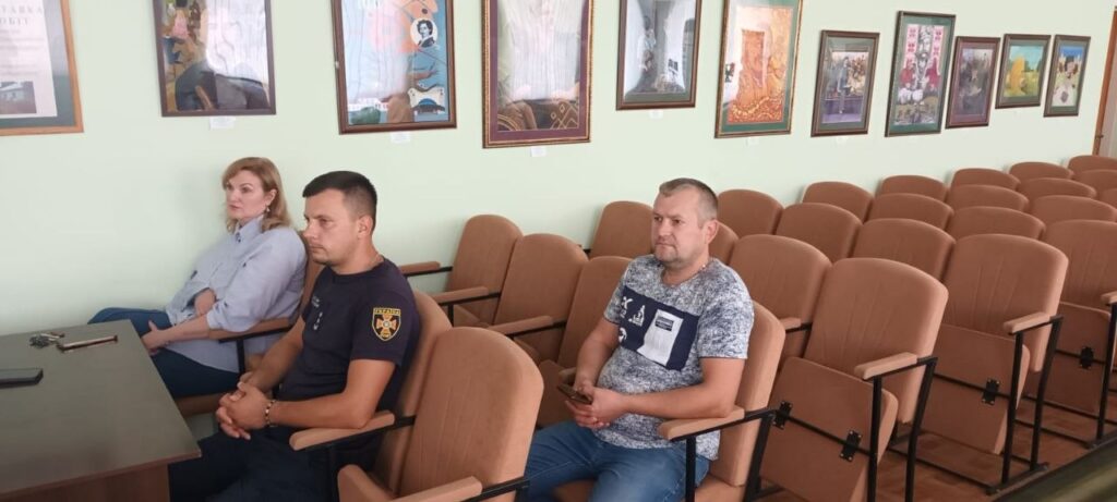 Три людини сидять на кріслах у залі з рядами порожніх місць та картинками на стіні.