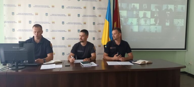Три чоловіки в формі сидять за столом під час відеоконференції з проєкцією учасників на екрані та прапорами на тлі.