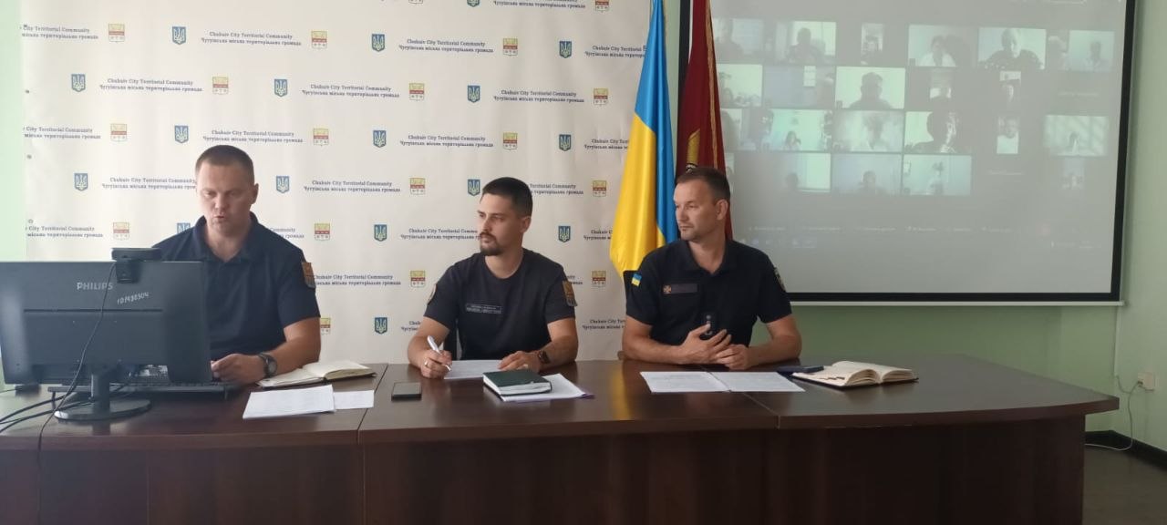 Три чоловіки в формі сидять за столом під час відеоконференції з проєкцією учасників на екрані та прапорами на тлі.