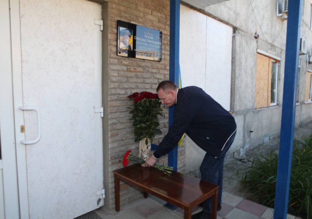 Чоловік у темному одязі кладе букет червоних троянд на дерев'яний столик біля входу з меморіальною дошкою.