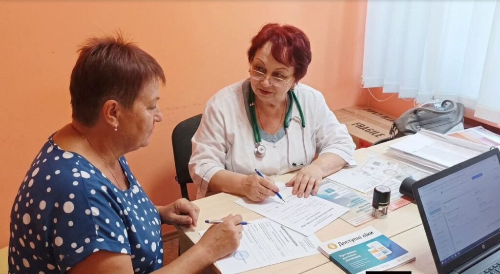 Лікарка зі стетоскопом на шиї консультує пацієнтку за столом, де лежать документи та буклет «Доступні ліки».