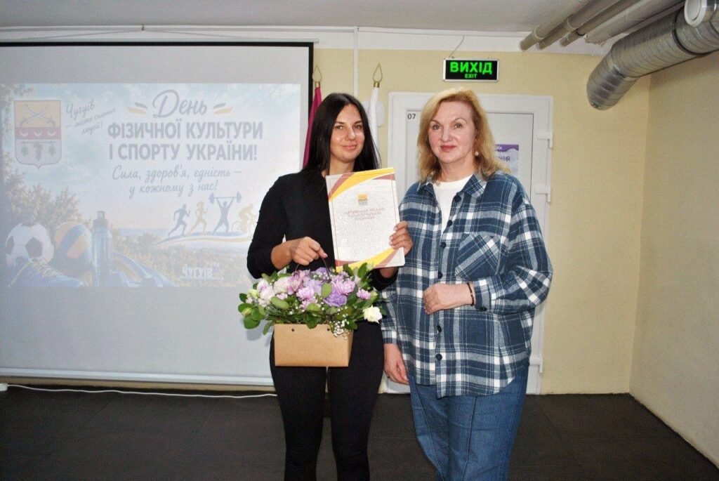 Дві жінки тримають квіти та сертифікат на тлі банера з написом «День фізичної культури і спорту України».