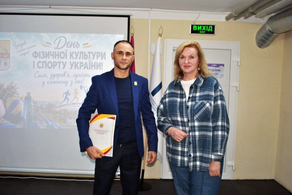 Чоловік у синьому костюмі тримає нагороду, стоячи поруч із жінкою в клітинковій сорочці на тлі банера «День фізичної культури і спорту України».