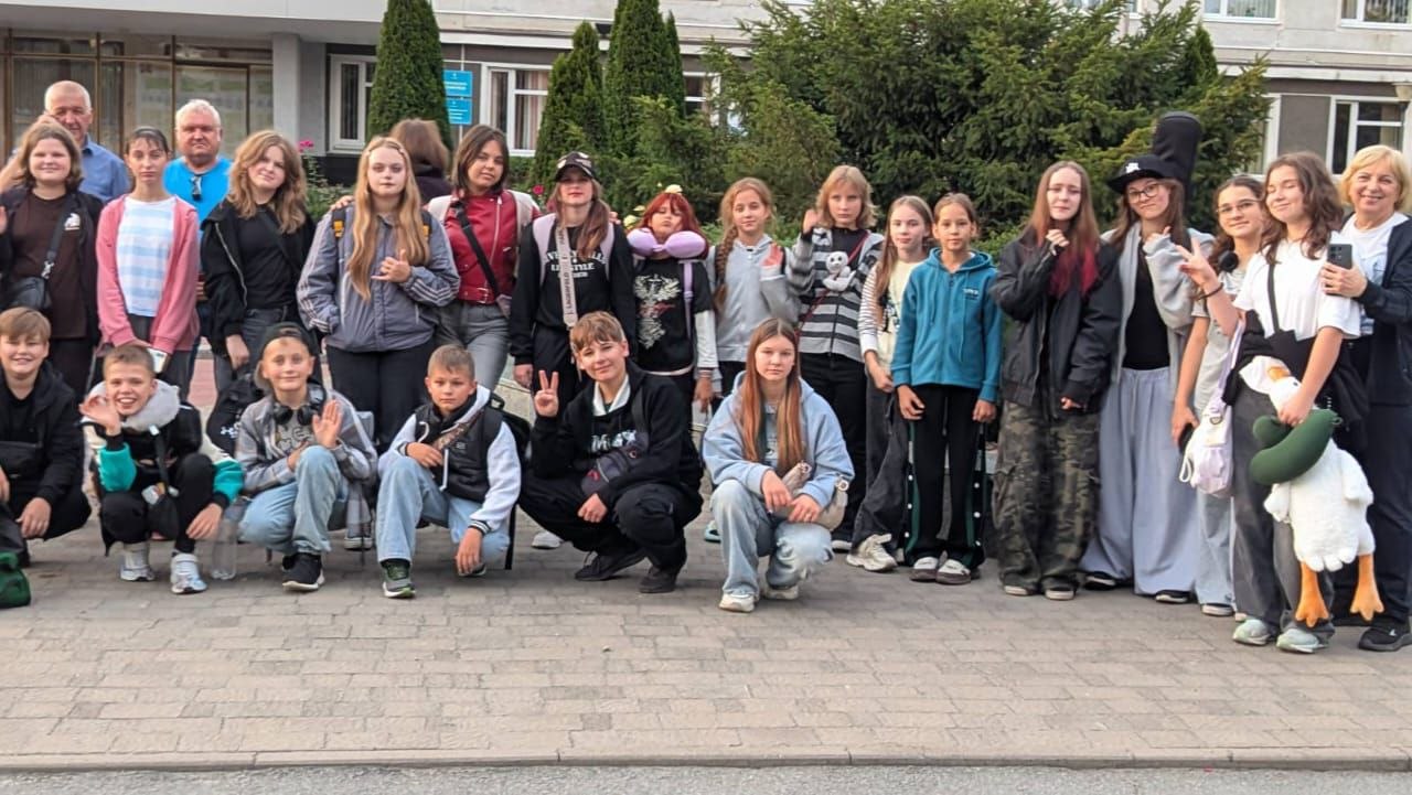 Група дітей та дорослих робить спільне фото на вулиці, де одна дівчина тримає велику іграшку качку, а інша одягнена в костюм із високою циліндричною шапкою.