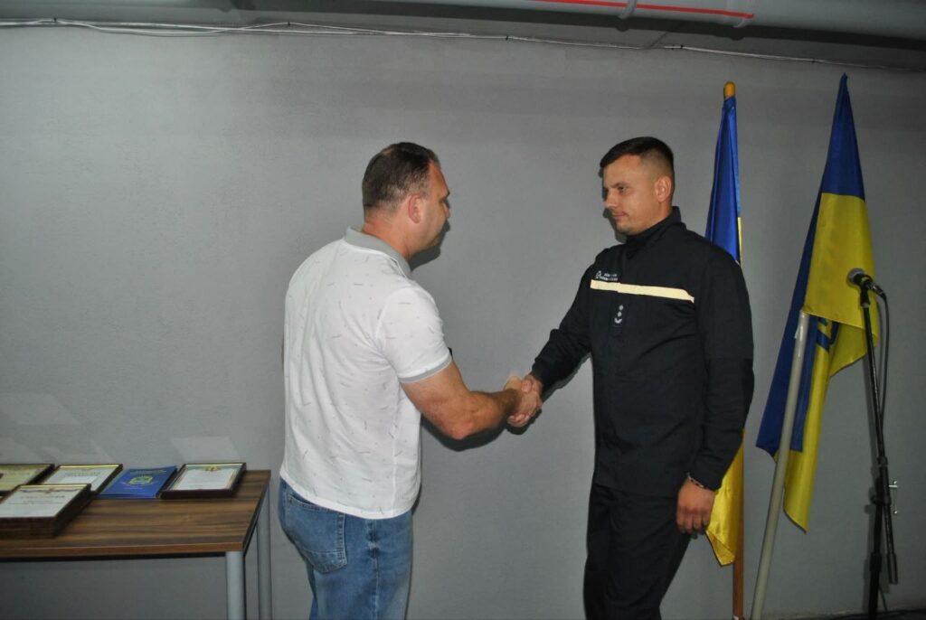 Два чоловіки поцілувалися в долоню на тлі прапорів ЄС та України, поруч стоїть стіл з нагородами.