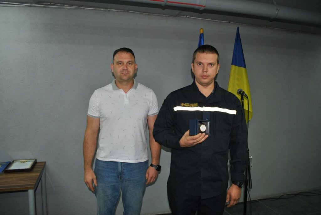 Двоє чоловіків, один у формі рятувальника, тримає нагороду в синьому футлярі на тлі українського прапора.