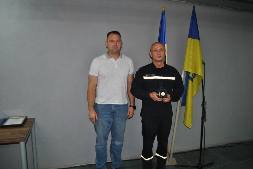 Два чоловіки стоять біля прапорів, один у формі тримає нагороду.