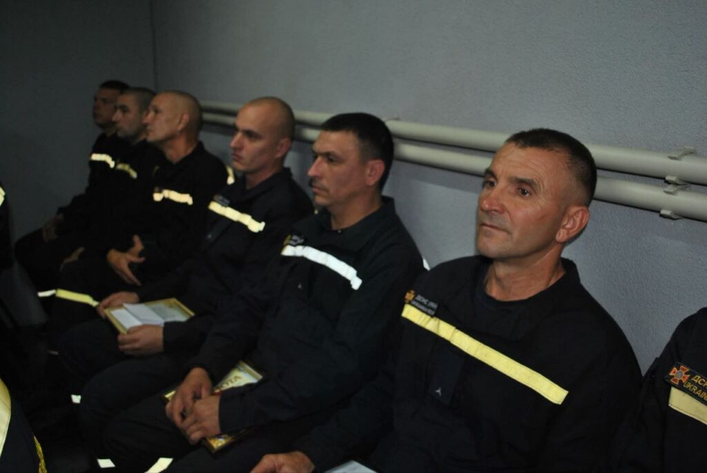 Група рятувальників у чорних робочих комбезах з жовтими відблискуючими смугами сидить у ряд, уважно слухаючи, а один з них тримає відкриту книгу.
