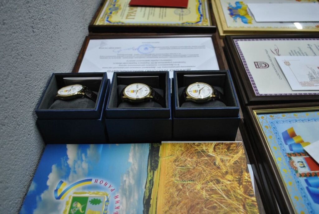 Золоті наручні годинники в чорних коробках, розташовані поруч із рамками з грамотами та фотокартками.
