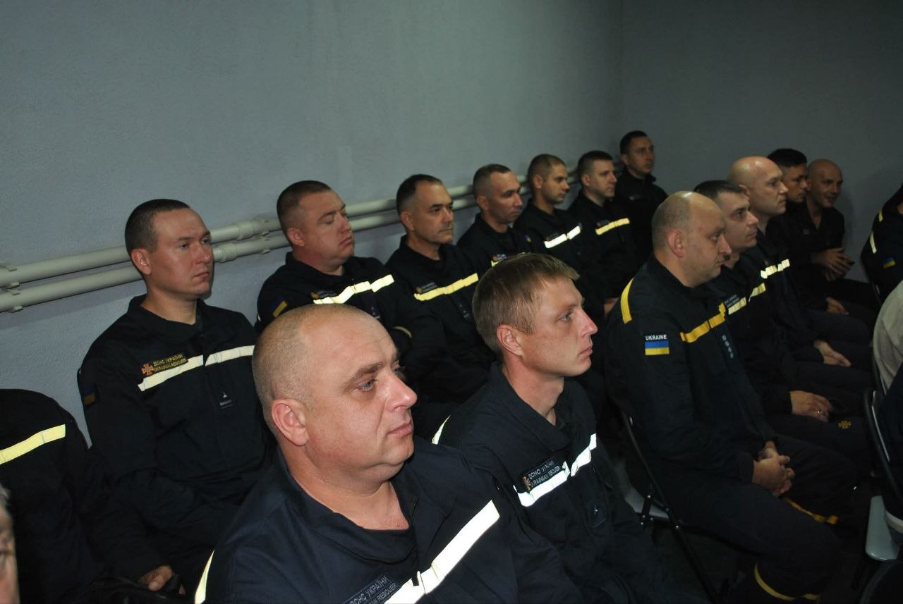 Група рятувальників у чорних формах з жовтими смугами та прапором України сидить рядами, уважно слухаючи.