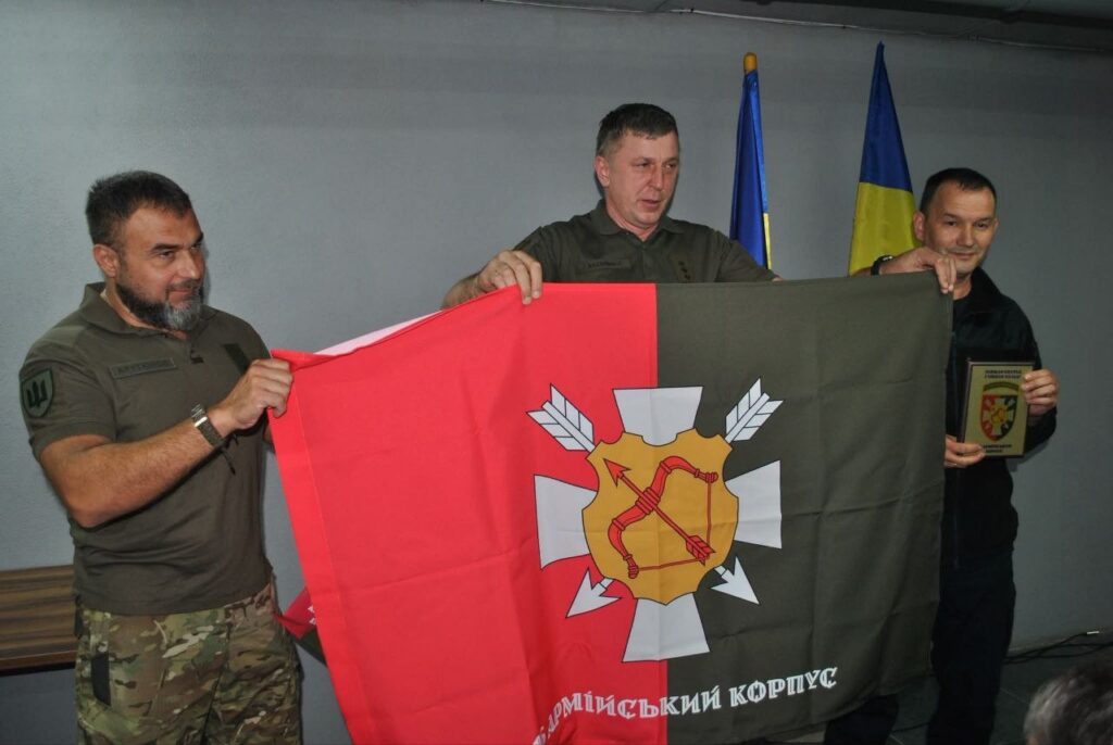 Військовослужбовці тримають прапор з написом «Армійський корпус» та емблемою з луком і стрілами.