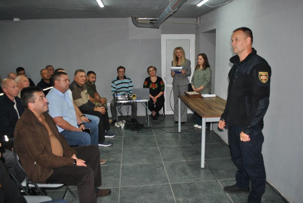 Чоловік у формі стоїть біля столу, звертаючись до групи людей, що сидять на стільцях, тоді як жінка в костюмі тримає документи.
