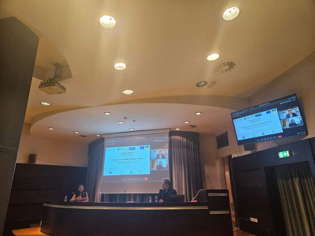 Учасники онлайн-конференції «Ukrainian Academy» сидять за столом у залі, дивлячись на екран з відеодзвінком.