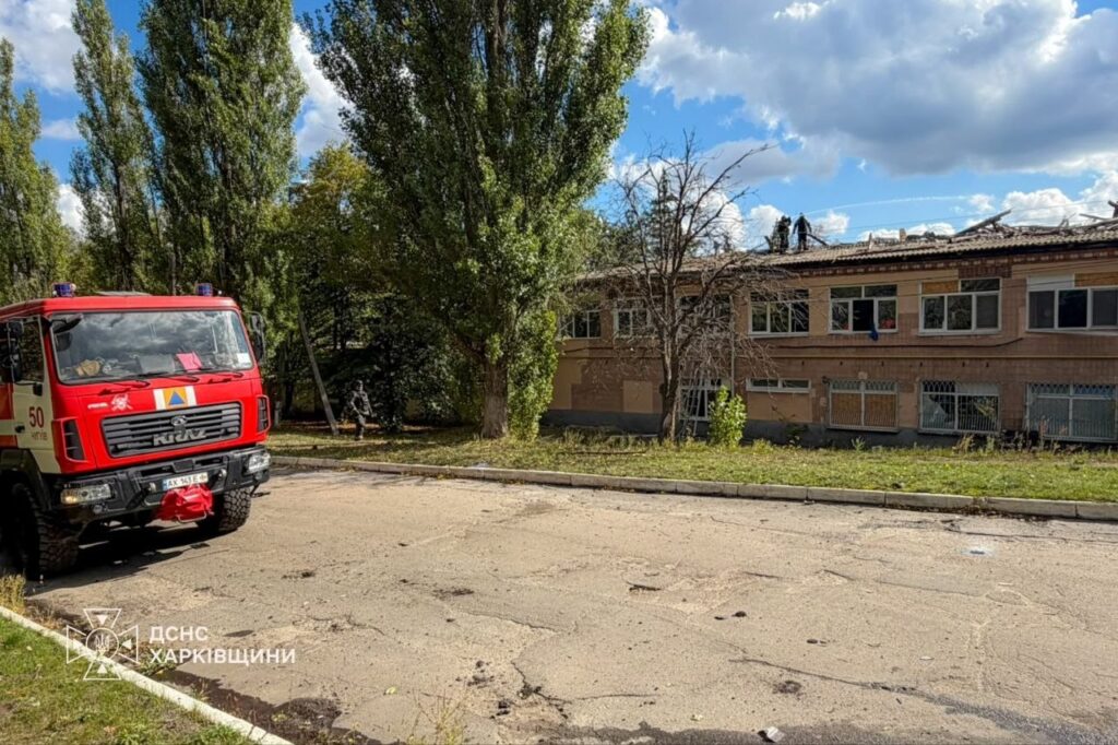 Пожежна машина ДСНС Харківщини стоїть біля будівлі з пошкодженою дахом, де працюють рятувальники.
