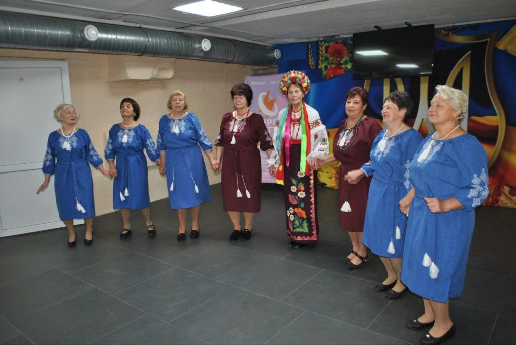 Група жінок у традиційних синіх та бордових сукнях з вишивкою, а також в українському народному вбранні з вінком, тримаються за руки в залі.