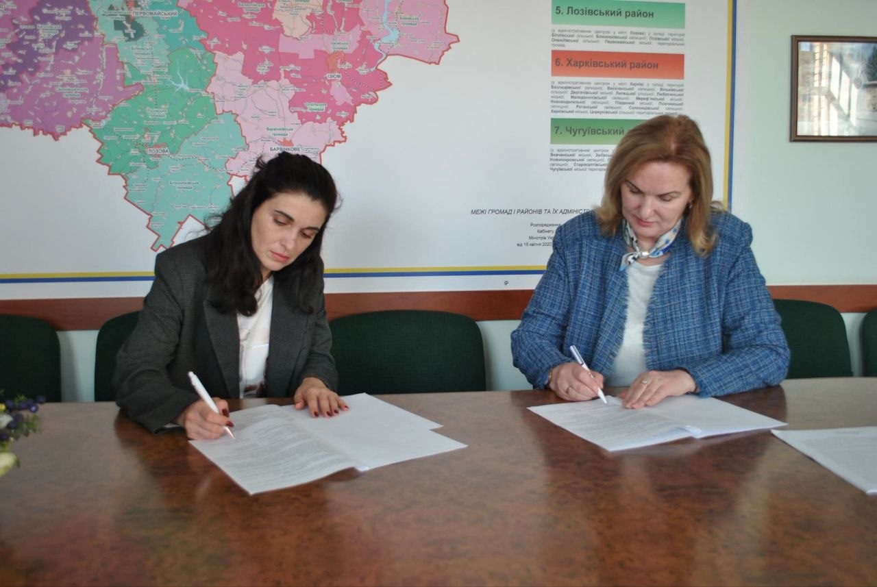 Дві жінки підписують документи за столом на тлі адміністративної карти.