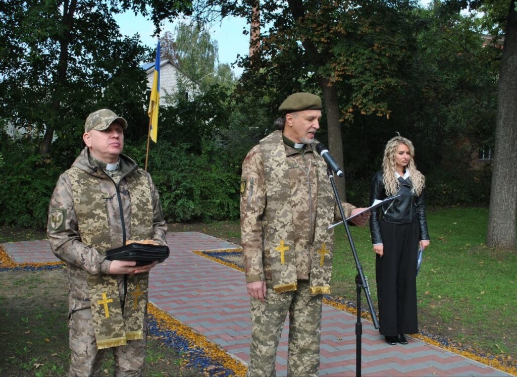 Духовний служитель у камуфляжі з хрестами на одязі читає молитву в мікрофон, а поруч стоїть інший священнослужитель, що тримає книгу, та жінка на тлі українського прапора.
