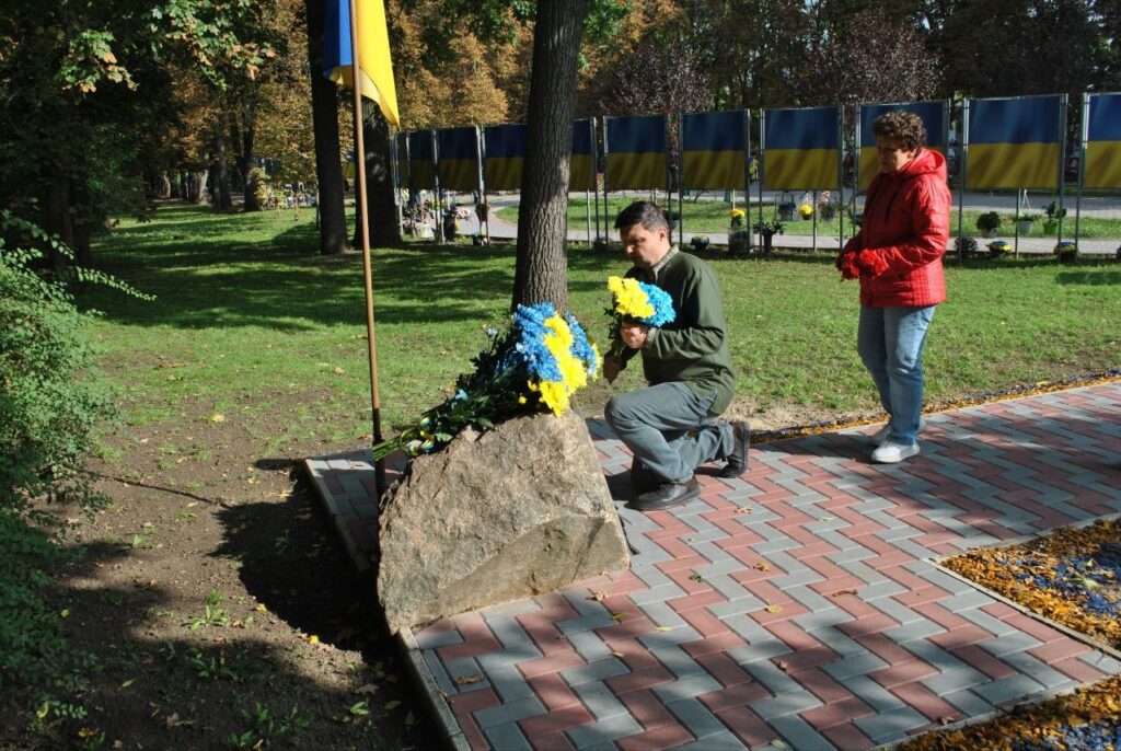 Людина в червоному куртку стоїть поруч із чоловіком, який кладе синьо-жовті квіти до каменю на тлі українських прапорів.