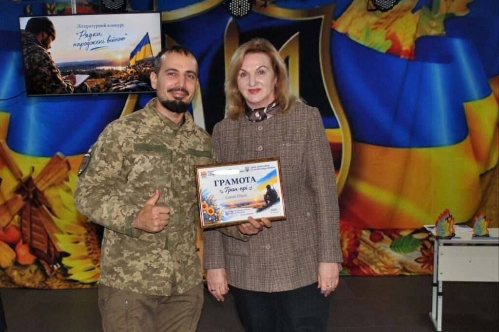 Військовослужбовець у камуфляжі та жінка у костюмі тримають нагороду з написом «ГРАМОТА Гран-прі» на тлі банера літературного конкурсу «Рядки, народжені війною».