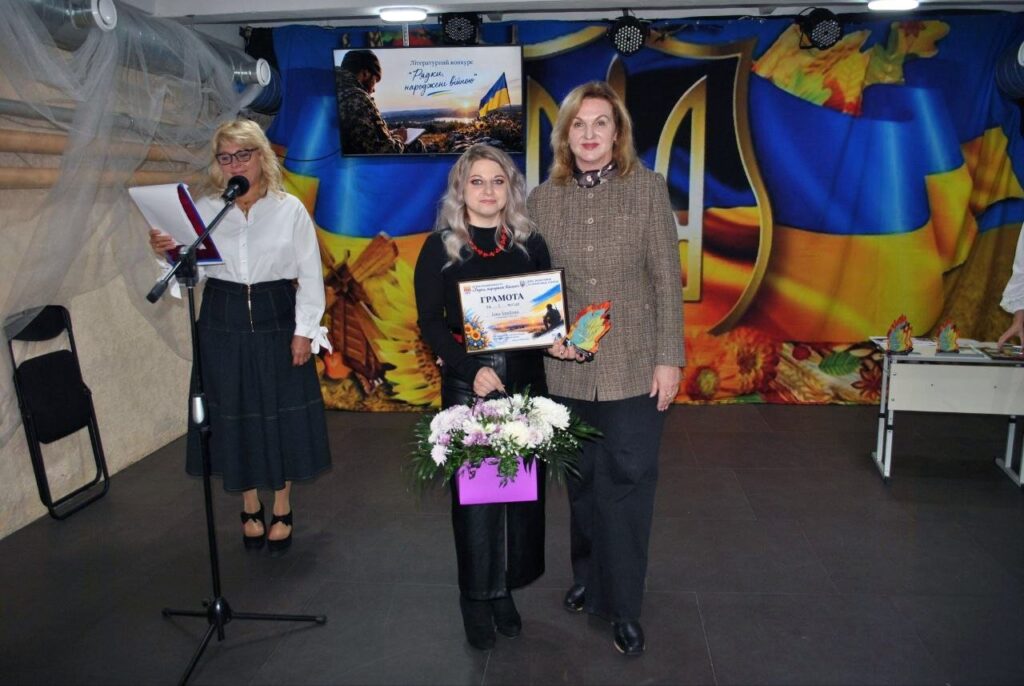 Жінка тримає грамоту та букет квітів, стоячи поруч з іншою жінкою на тлі банера літературного конкурсу «Радки народжені бійком».