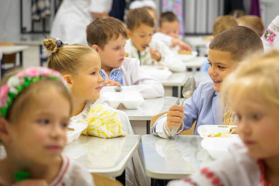 Діти в білих сорочках сидять за шкільними партами та їдять з тарілок.