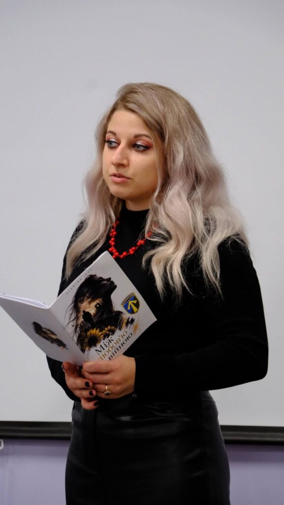 Жінка з довгим світлим волоссям у чорному одязі тримає відкриту книгу з написом «Міфологія» та зображенням сокола.